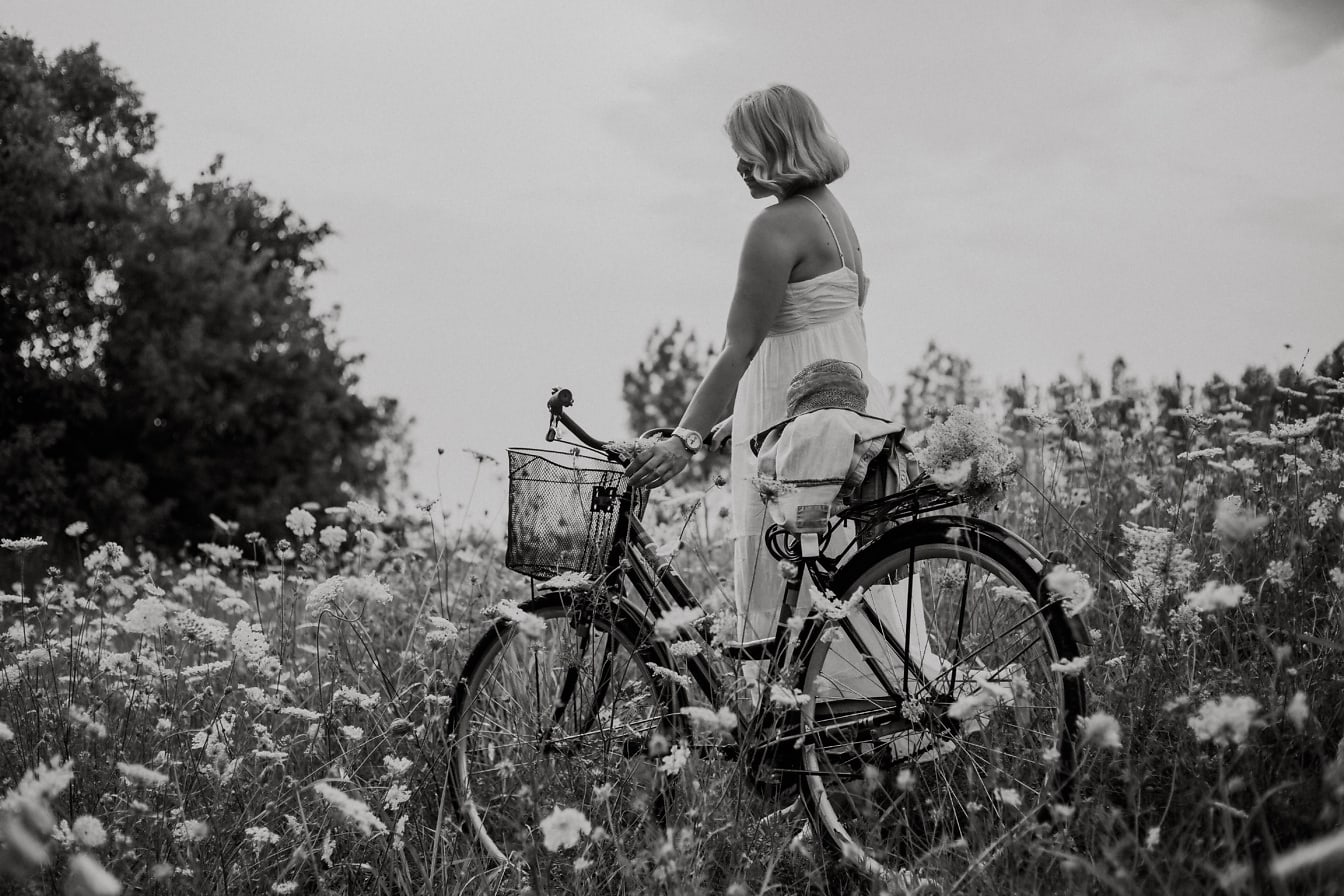 Wanita berdiri dengan sepeda di padang rumput dengan bunga foto hitam putih