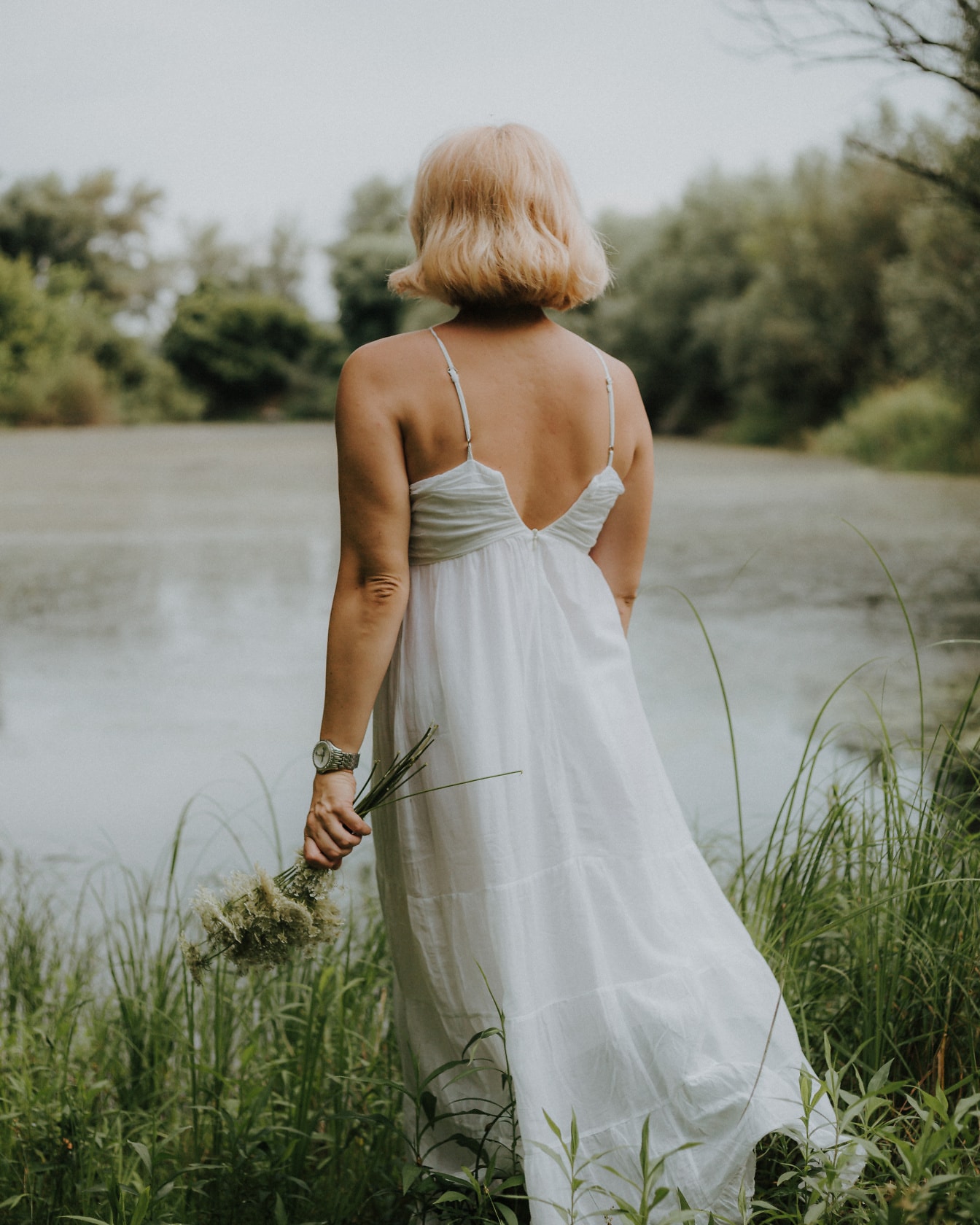 Žena v bielych šatách bez chrbta drží kvety vo vysokej tráve na brehu jazera