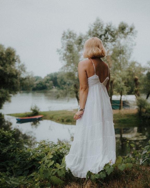 Blonde woman in a white backless dress standing by lake water
