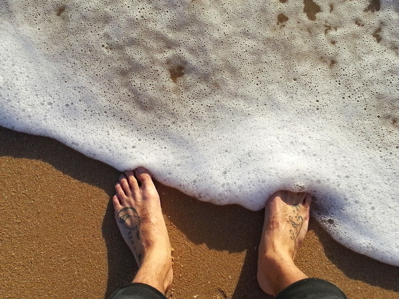 Person mit Tätowierungen an den Füßen, die auf nassem Sand in Schaumwasser stehen