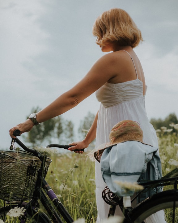 Frau in einem weißen rückenfreien Kleid mit Strohhut auf einem Fahrrad