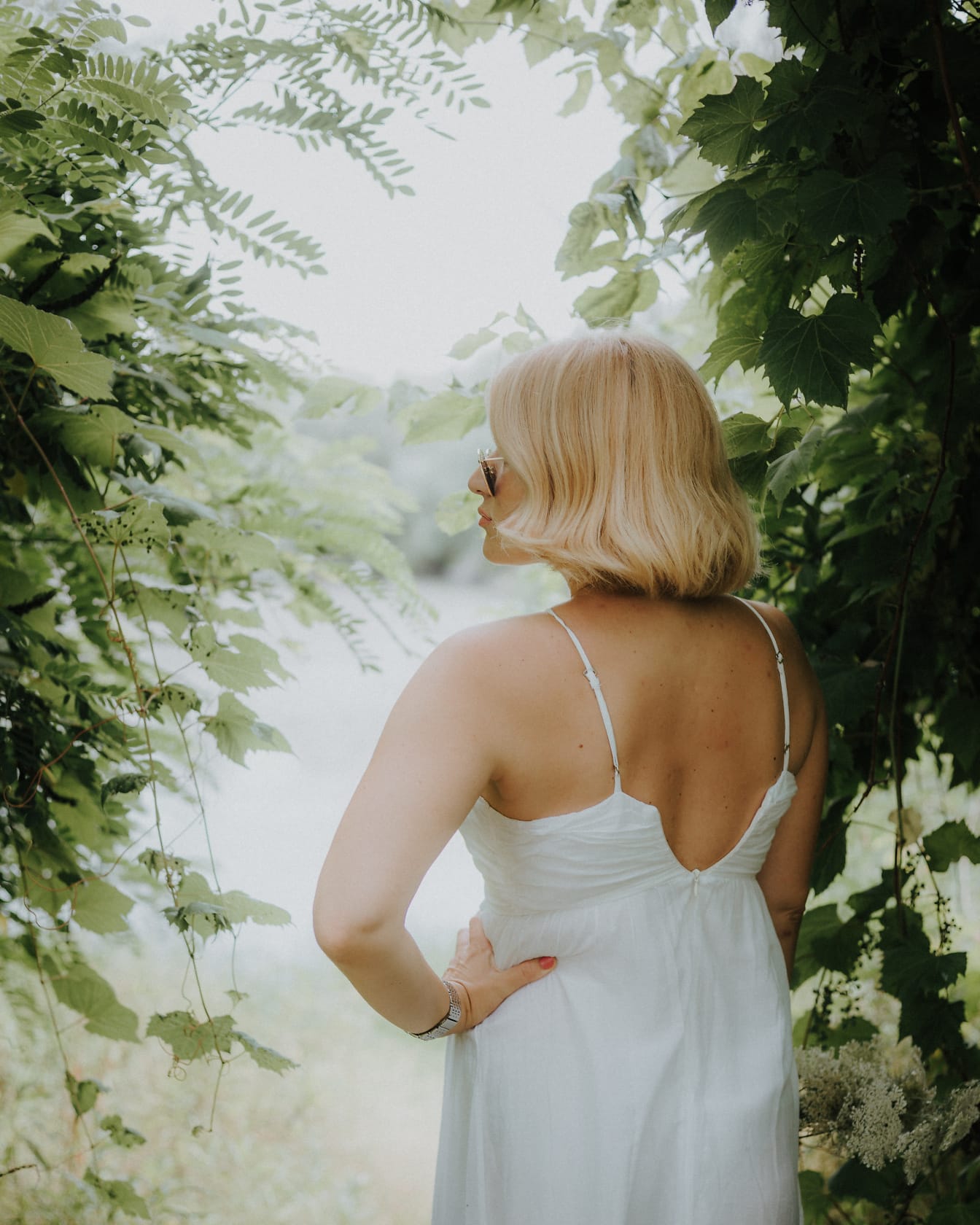 Žena u bijeloj vjenčanici bez leđa stoji među zelenim lišćem
