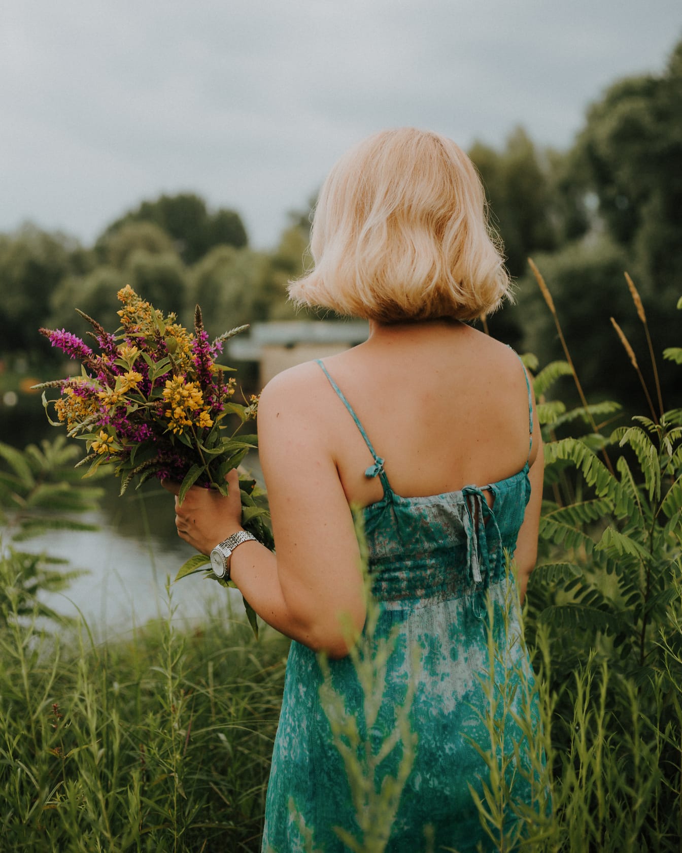 Kobieta odwrócona plecami w satynowej sukience bez pleców trzymająca bukiet kwiatów na polu