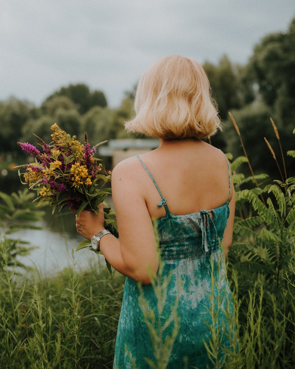 Žena otočená zády v saténových šatech s odhalenými zády držící kytici květin na poli