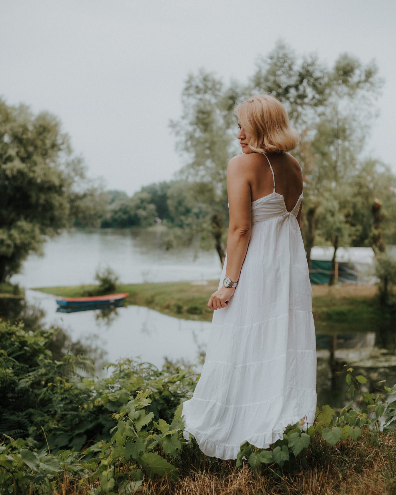 白い背中の開いたウェディングドレスを着た恥ずかしがり屋の花嫁が川のそばに立っています