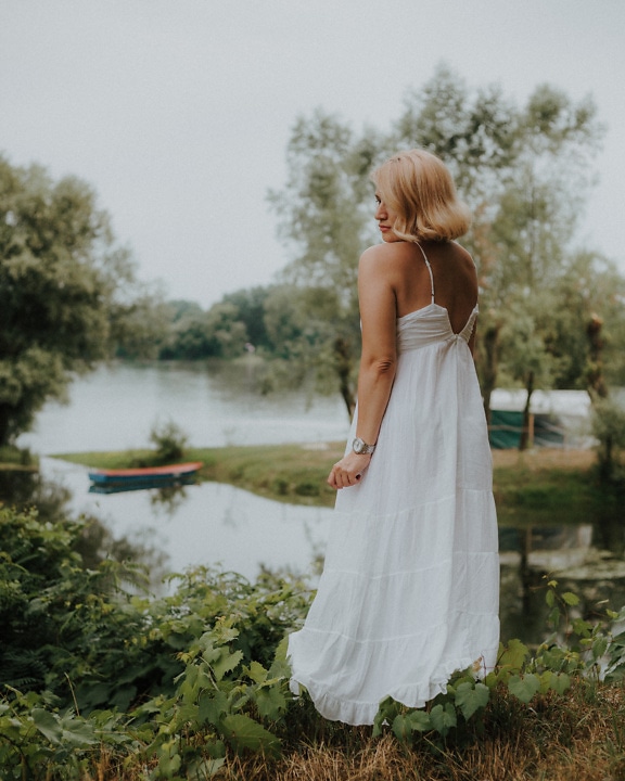 A shy bride in a white backless wedding dress stands by the river