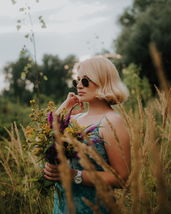 Blond žena s okuliarmi držiaca kvety vo vysokej tráve