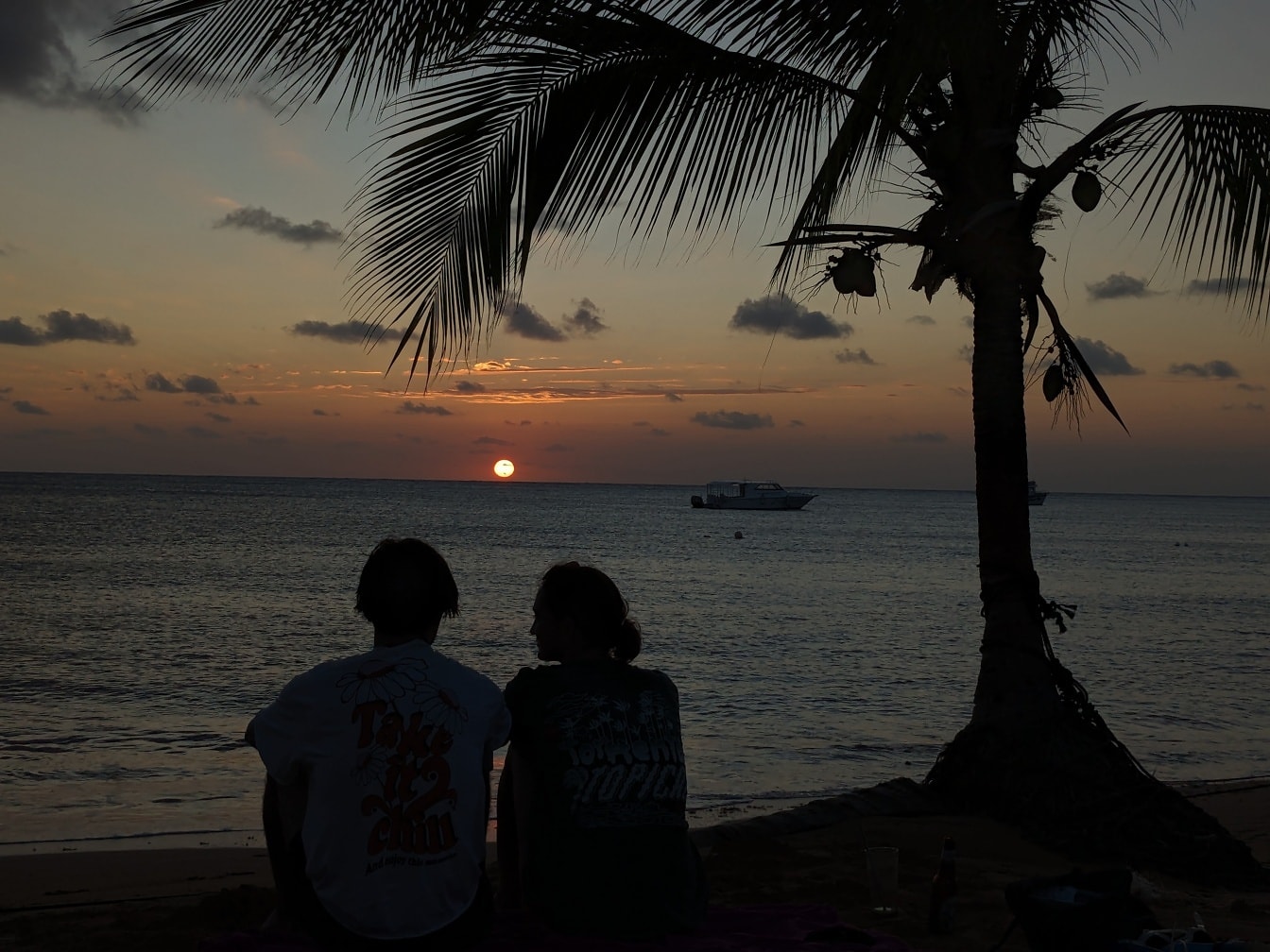 Romantikus pár sziluettje, akik a tengerparton ülnek pálmafa alatt, és élvezik a naplementét