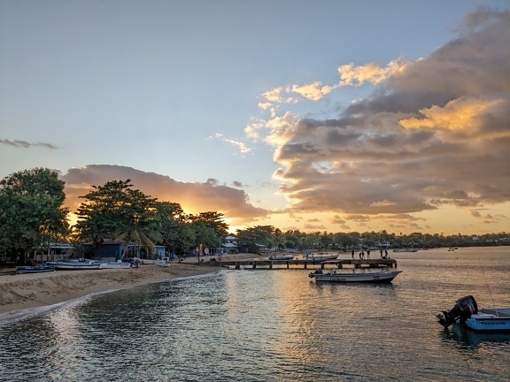 Tropisk strand med båter og brygge i bakgrunnen ved soloppgang