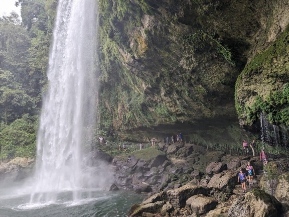 Touristen, die auf einem Fußgängerweg unter dem Wasserfall spazieren gehen