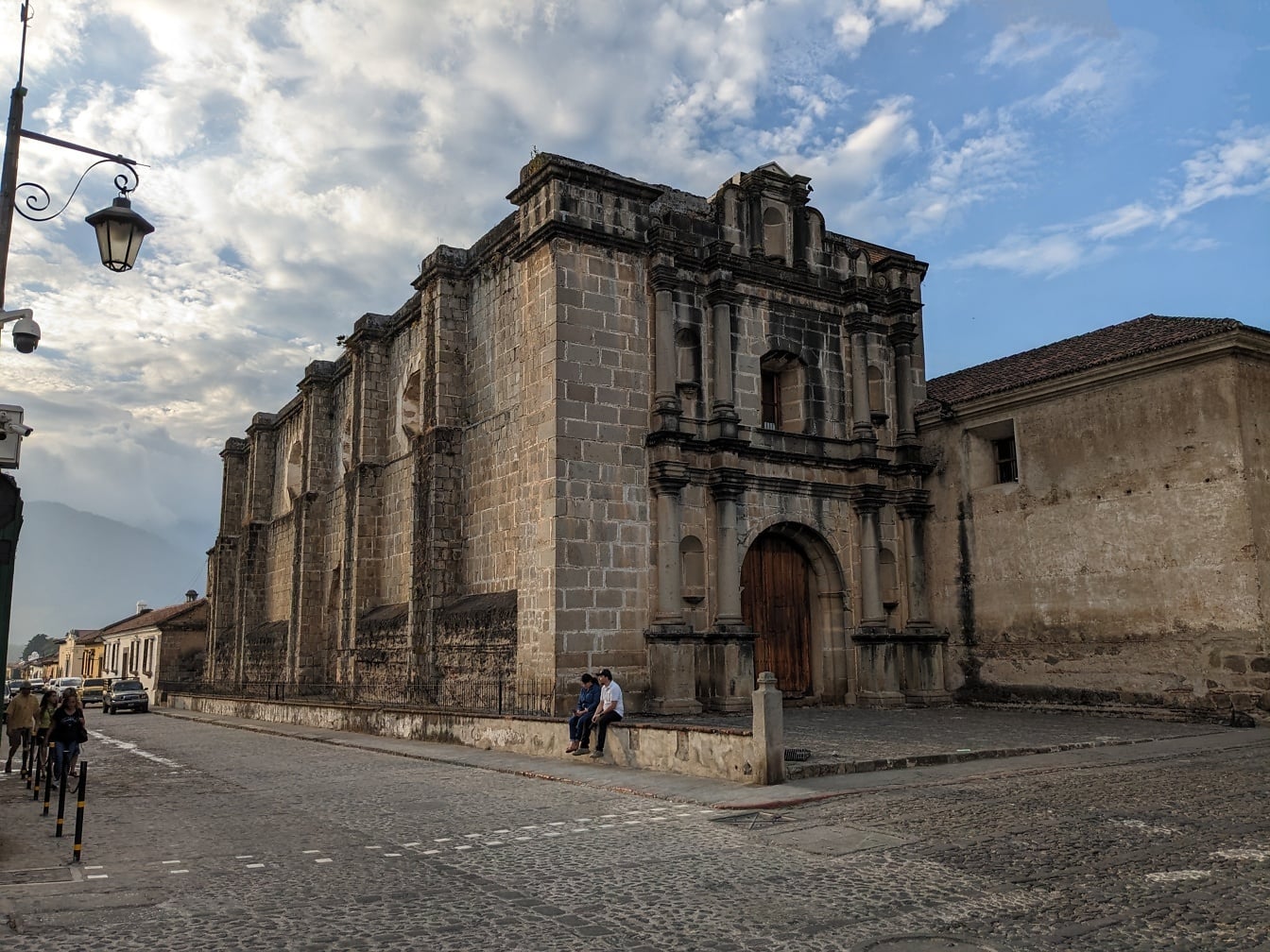 Ruinen eines Steingebäudes in Antigua, Guatemala, mit ein paar Leuten, die auf einer Straße sitzen