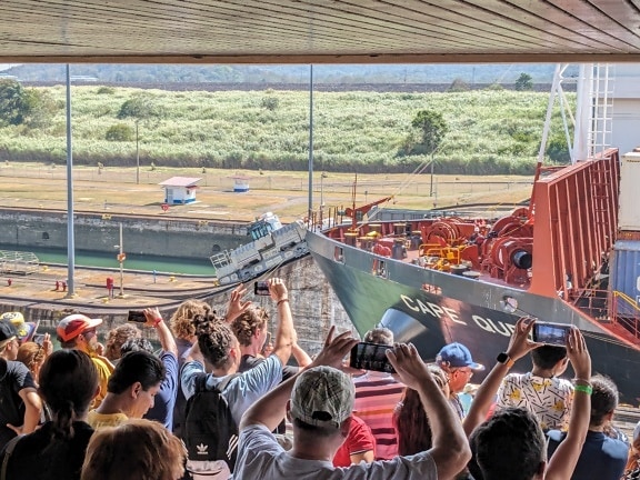 People watch and take photos with their cell phones of a large ship in the Panama Canal
