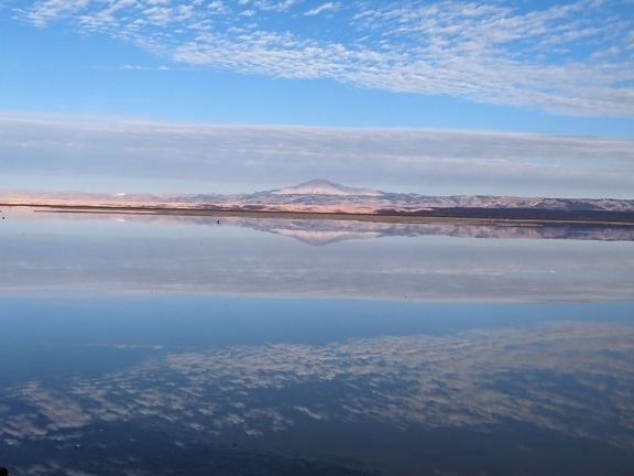 Reflet d’un ciel bleu avec des nuages et une montagne au loin dans un lac salé