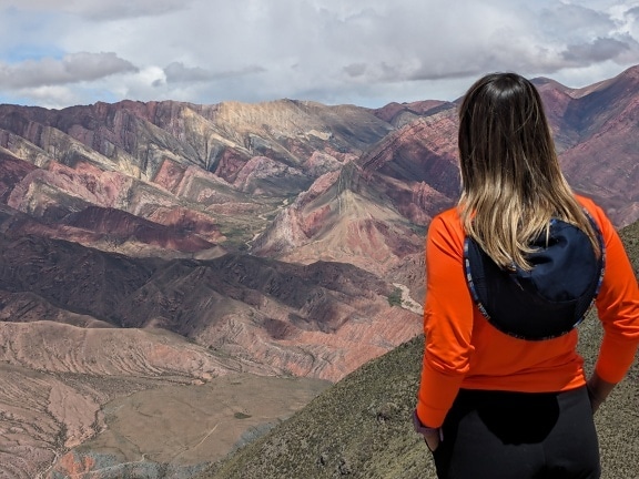 Odwrócona plecami kobieta z kapeluszem na plecach stoi na szczycie góry i podziwia panoramę doliny