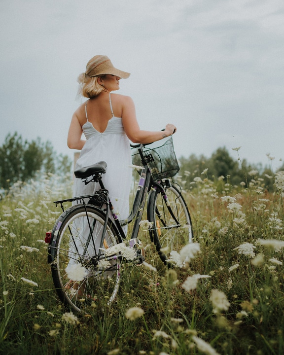 Woman in a white backless dress and hat with a black bicycle in a field of wildflowers