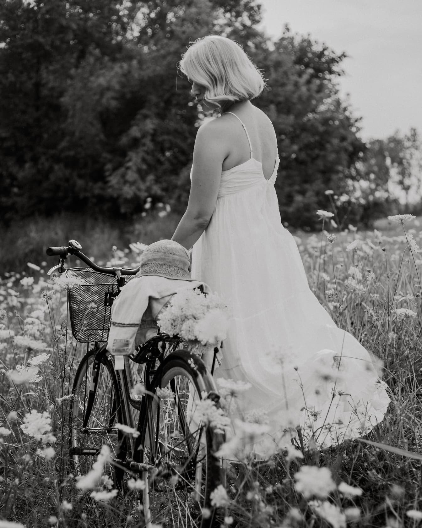 Mireasa țării într-o rochie populară albă, fără spate, cu o bicicletă într-un câmp de flori, fotografie alb-negru