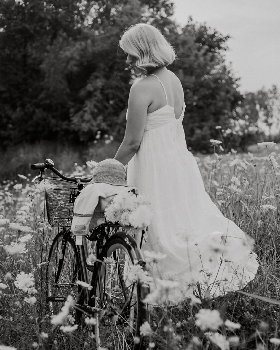꽃밭에서 자전거와 함께 등이 없는 흰색 민속 드레스를 입은 시골 신부, 흑백 사진