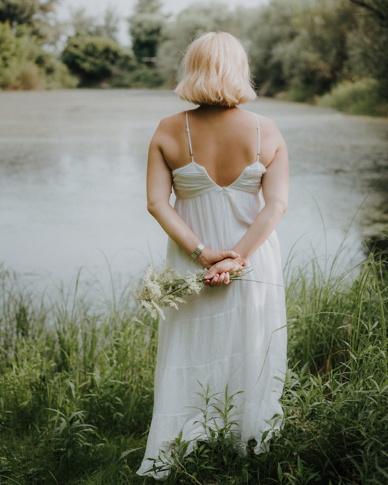 En ryggvänd brud som står i en vit klänning utan rygg vid sjön och håller blommor i sina händer