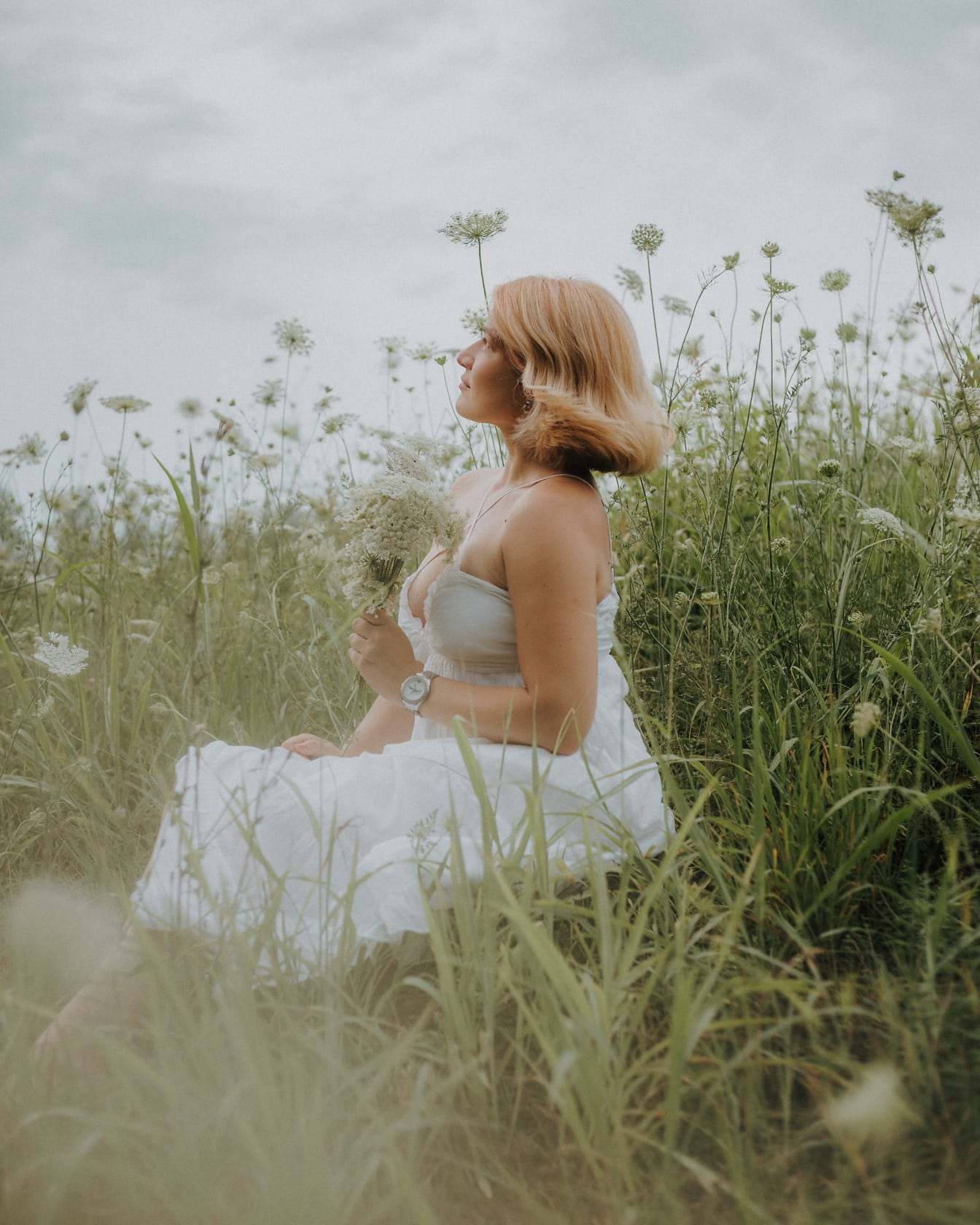 背の高い草の野原に座っている白い民俗のウェディングドレスを着たかわいい国の花嫁