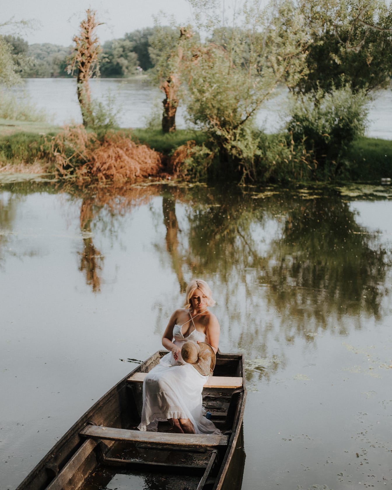 Ženská mladá žena ve venkovských bílých šatech sedí v dřevěné lodi na jezeře
