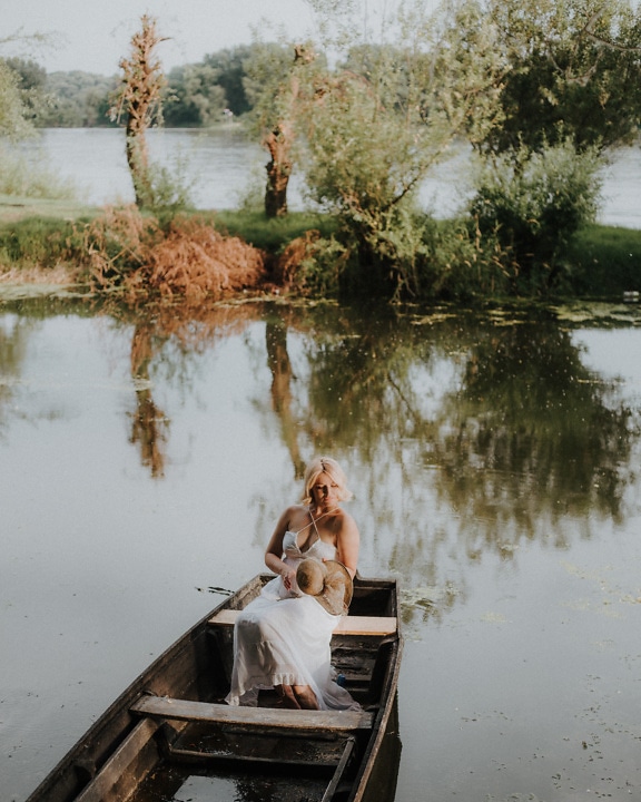 Người phụ nữ trẻ nữ tính trong chiếc váy trắng đồng quê ngồi trên một chiếc thuyền gỗ trên hồ