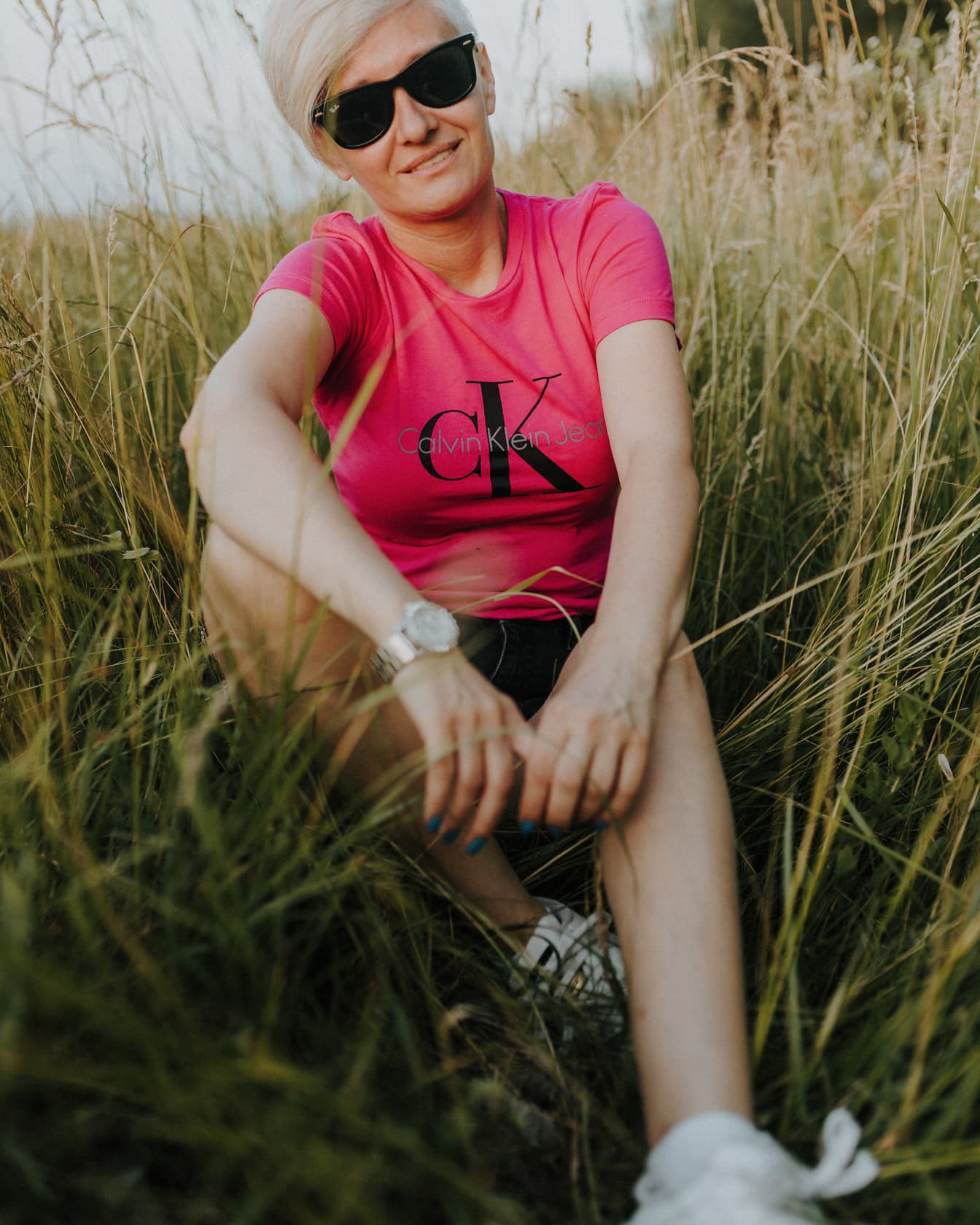 Usmievavá blondínka s krátkymi vlasmi so slnečnými okuliarmi sediaca vo vysokej tráve