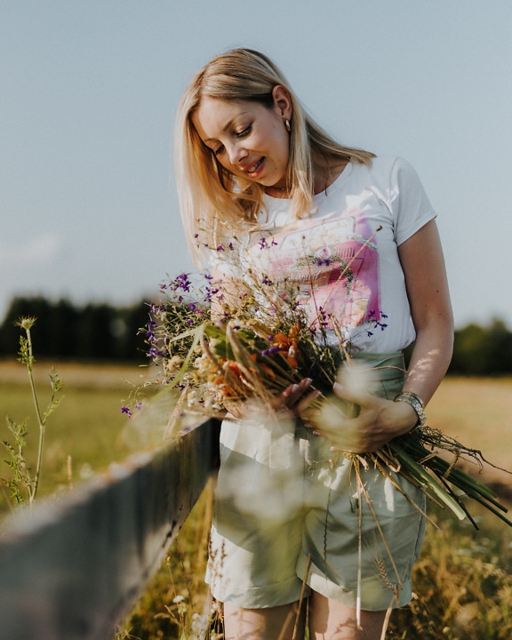 Smiling country young woman holding flowers in a field by farm fence