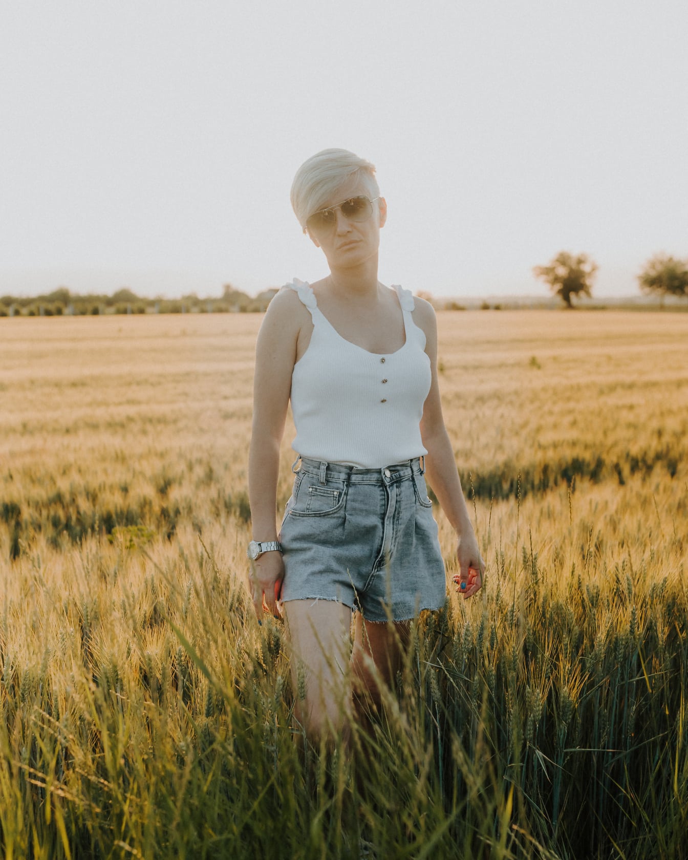 En korthåret kvinde i jeans, shorts og en hvid ærmeløs t-shirt står i en hvedemark;
