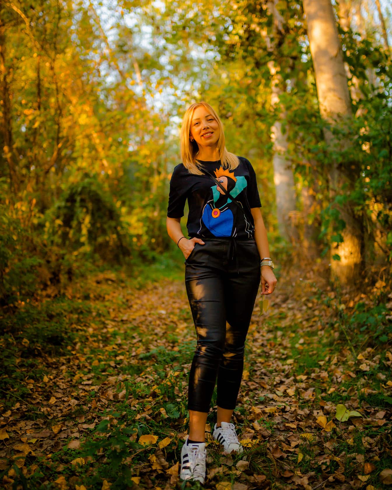 Kaunis hymyilevä blondi seisoo metsässä mustassa muodikkaassa asussa