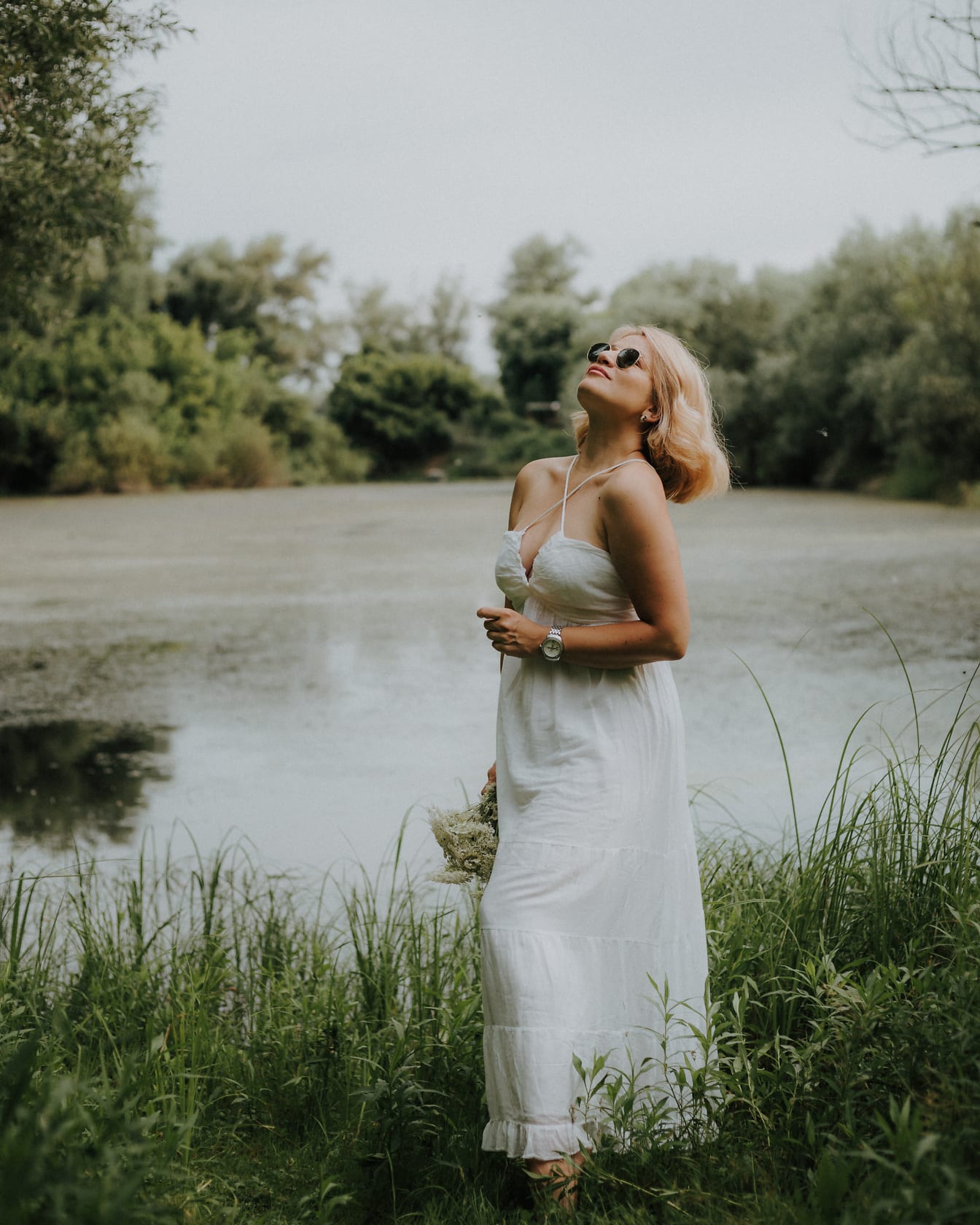 Wanita bangga berpose dalam gaun putih feminin berdiri di tepi danau