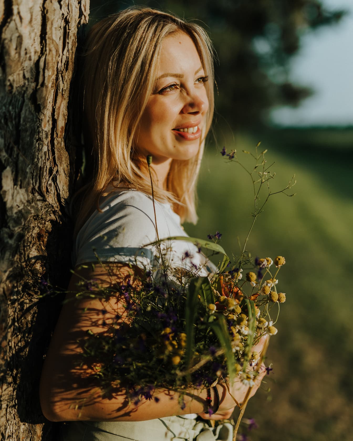 Portret lateral al unei femei zâmbitoare sprijinindu-se de un copac în timp ce ține flori