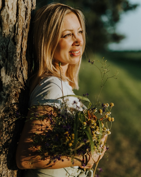 Страничен портрет на усмихната жена, облегната на дърво, докато държи цветя
