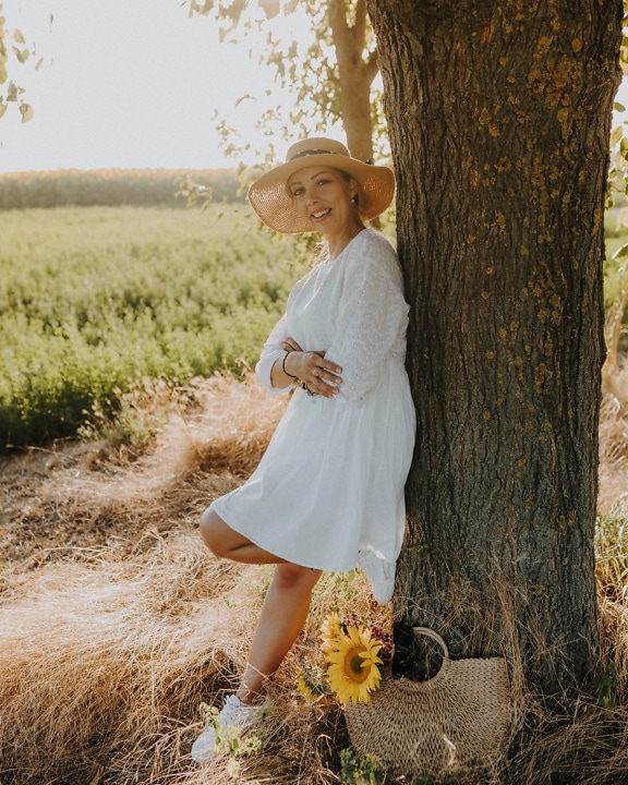 Smiling country young woman in white dress and straw-hat leaning against a tree in a field