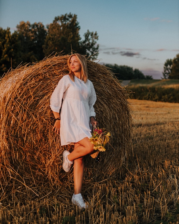 Country young woman in a white folk dress leaning on a bale of hay in wheat field