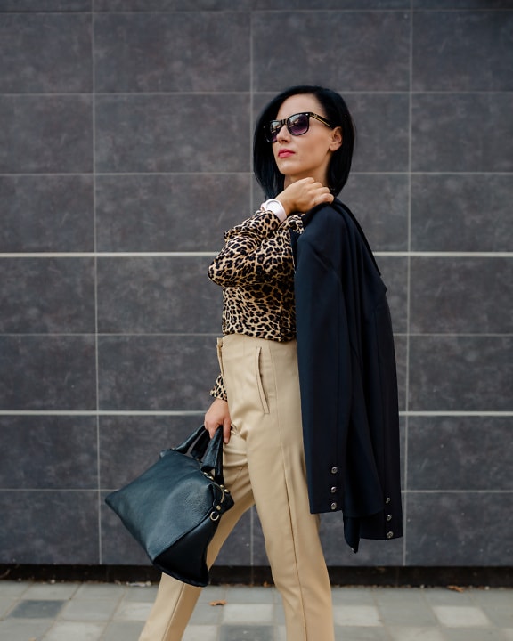 Selbstbewusste Geschäftsfrau in Hemd mit Leopardenmuster und beigefarbener Hose mit schwarzer Tasche in der Hand