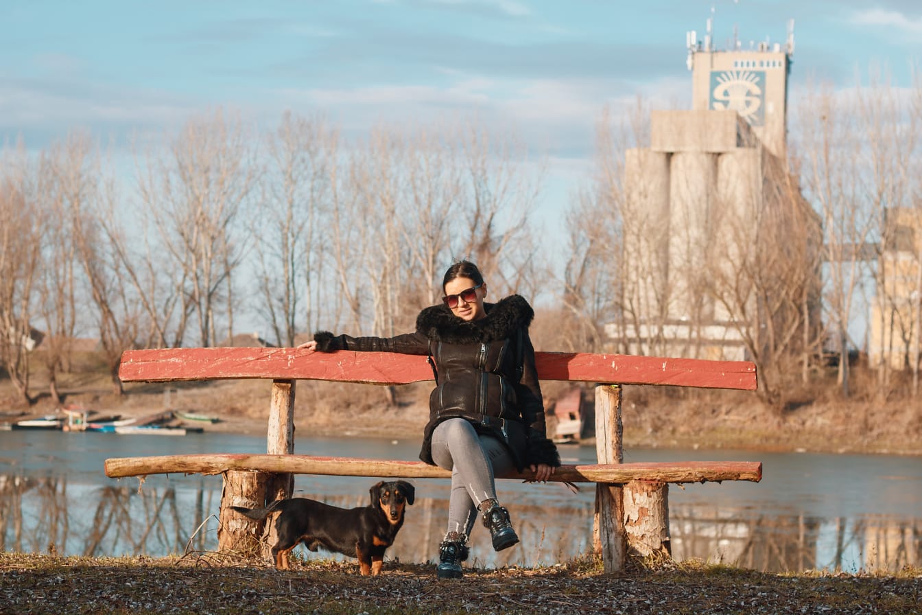 O femeie stă pe o bancă cu un câine și se bucură de o zi însorită de toamnă