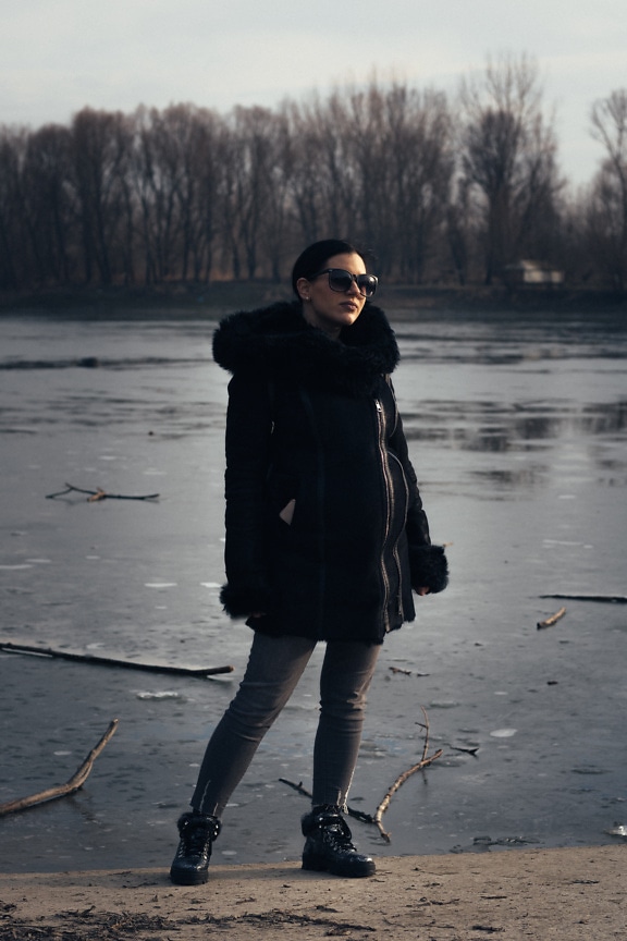 Good looking woman in black winter coat standing on a shore of frozen lake