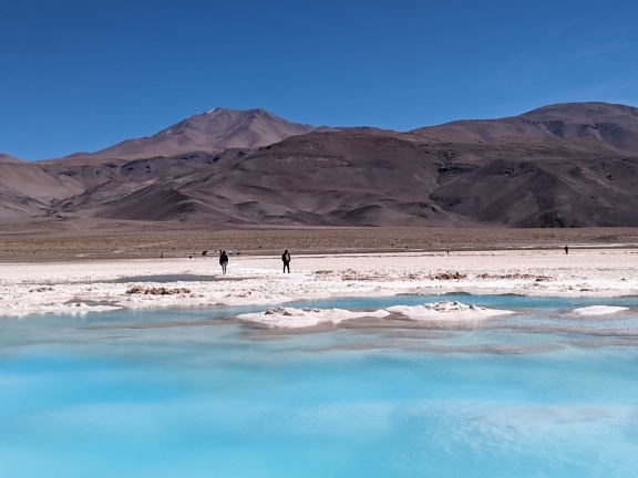 People standing on the salt crystals at beach of salt lake with azure color water in desert