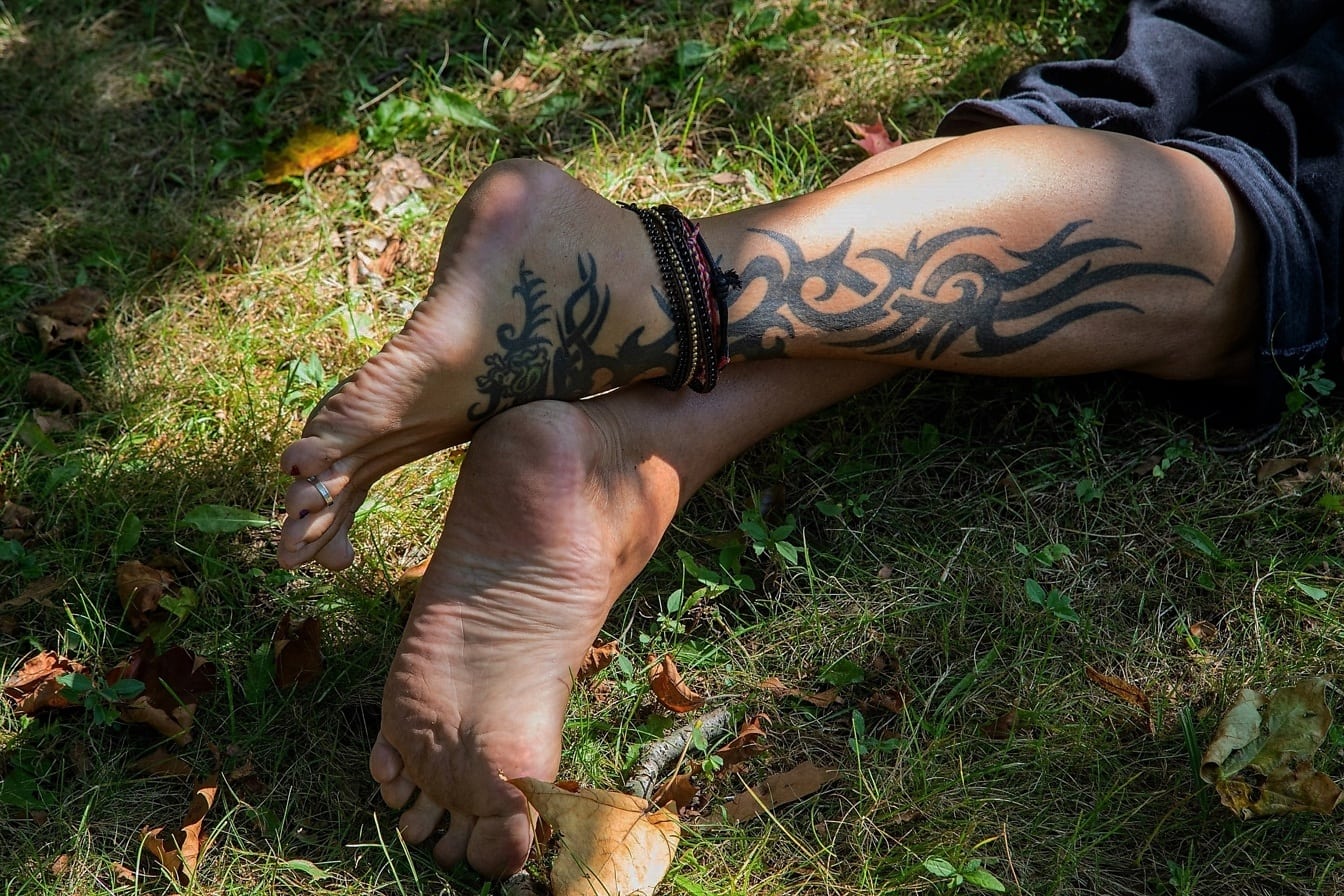 Gambe da uomo a piedi nudi con tatuaggi e braccialetti alla caviglia