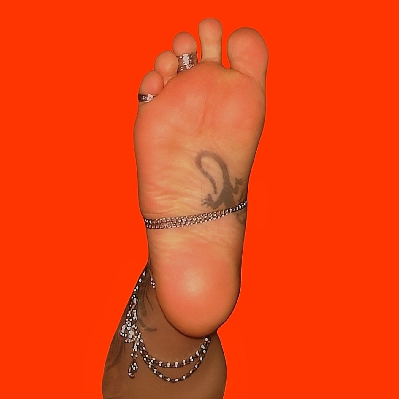 Tetované pánské bosé nohy s kotníkovými náramky a prsteny s oranžovým pozadím