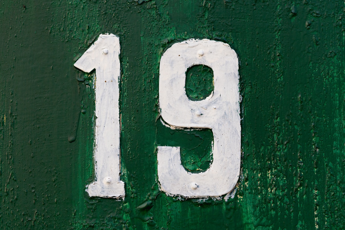 Kov číslo 19 lakovaný bílou barvou na tmavě zeleném kovovém povrchu