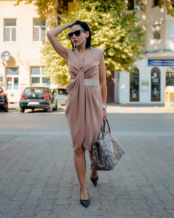 Eine schlanke Frau-Fotomodell in einem beigen Kleid und mit modischer Handtasche mit Schlangenprint geht auf einem Bürgersteig