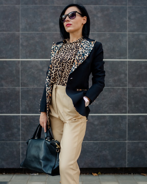 Hermosa mujer de negocios con traje con estampado de leopardo y pantalones brillantes posando sosteniendo un bolso de cuero negro