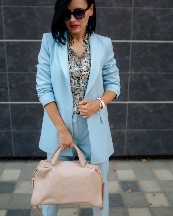 A slender young businesswoman in blue pants and a business suit holding a beige leather handbag