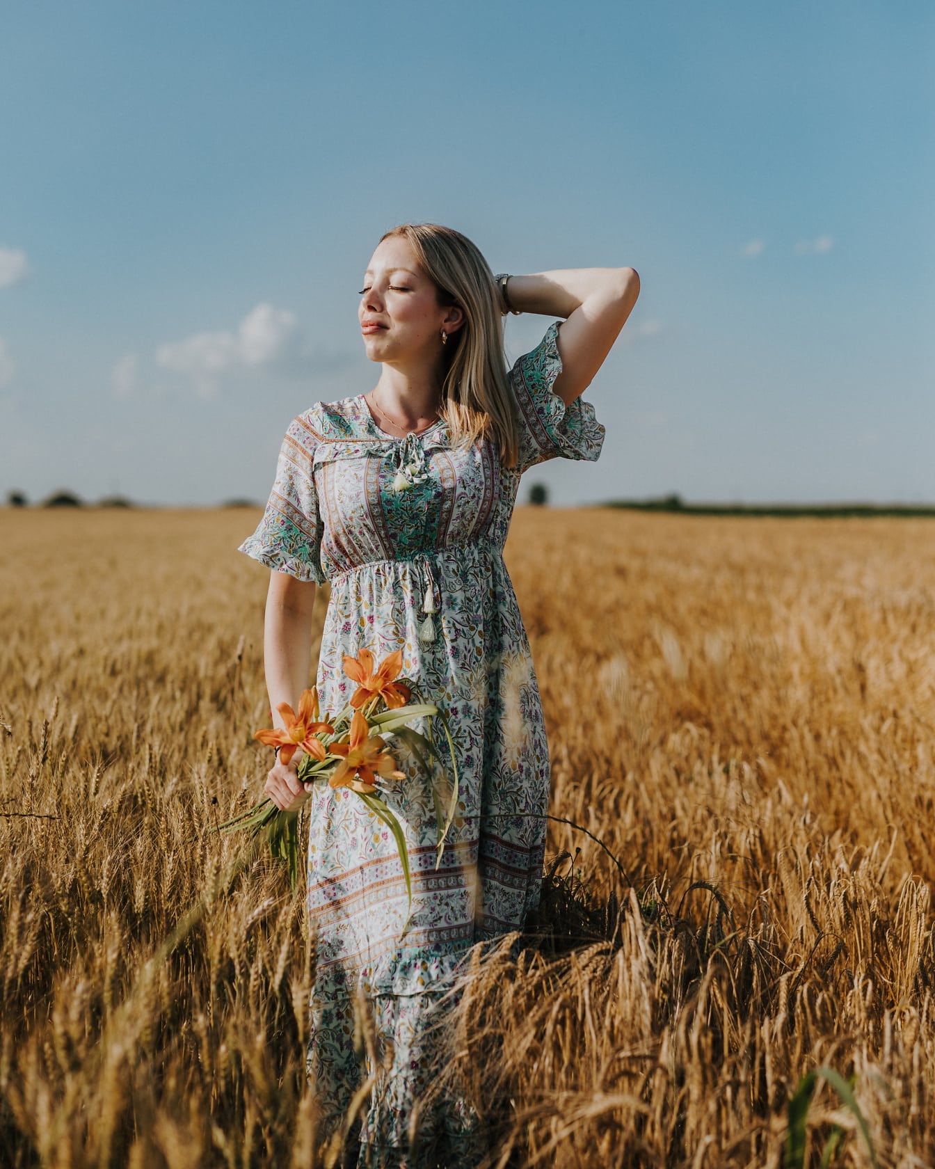 Porträtt av en fantastiskt vacker blondin i en lantlig stilklänning som rymmer blommor i ett vetefält på sommartid