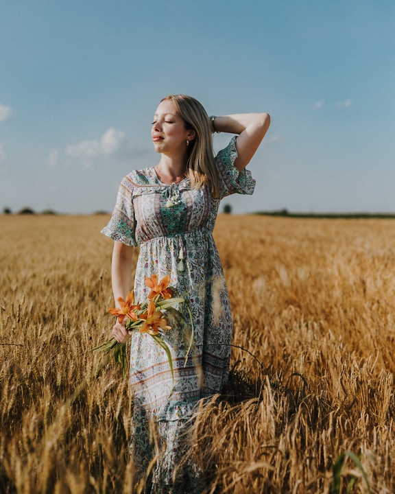 Portrait of a stunningly beautiful blonde in a country style dress holding flowers in a field of wheat at summer time