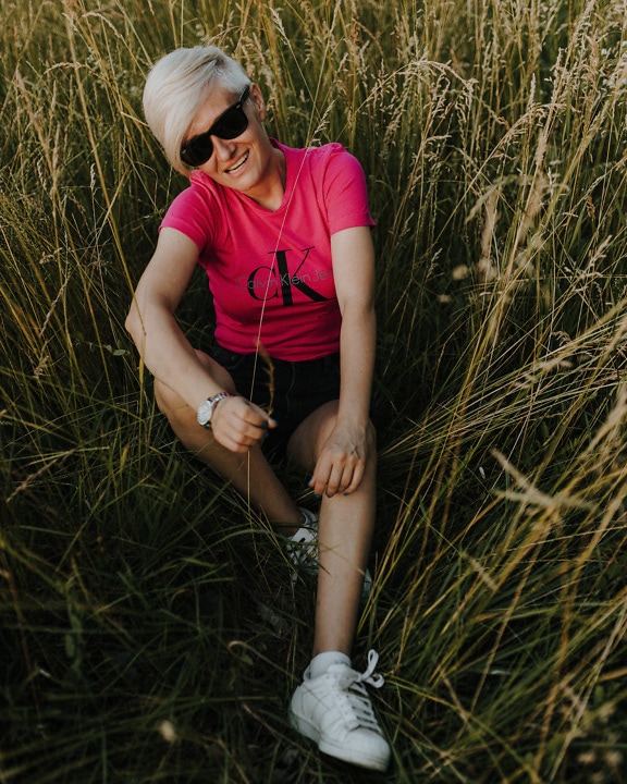 A young woman with short blonde hair sits in the tall grass and smiles