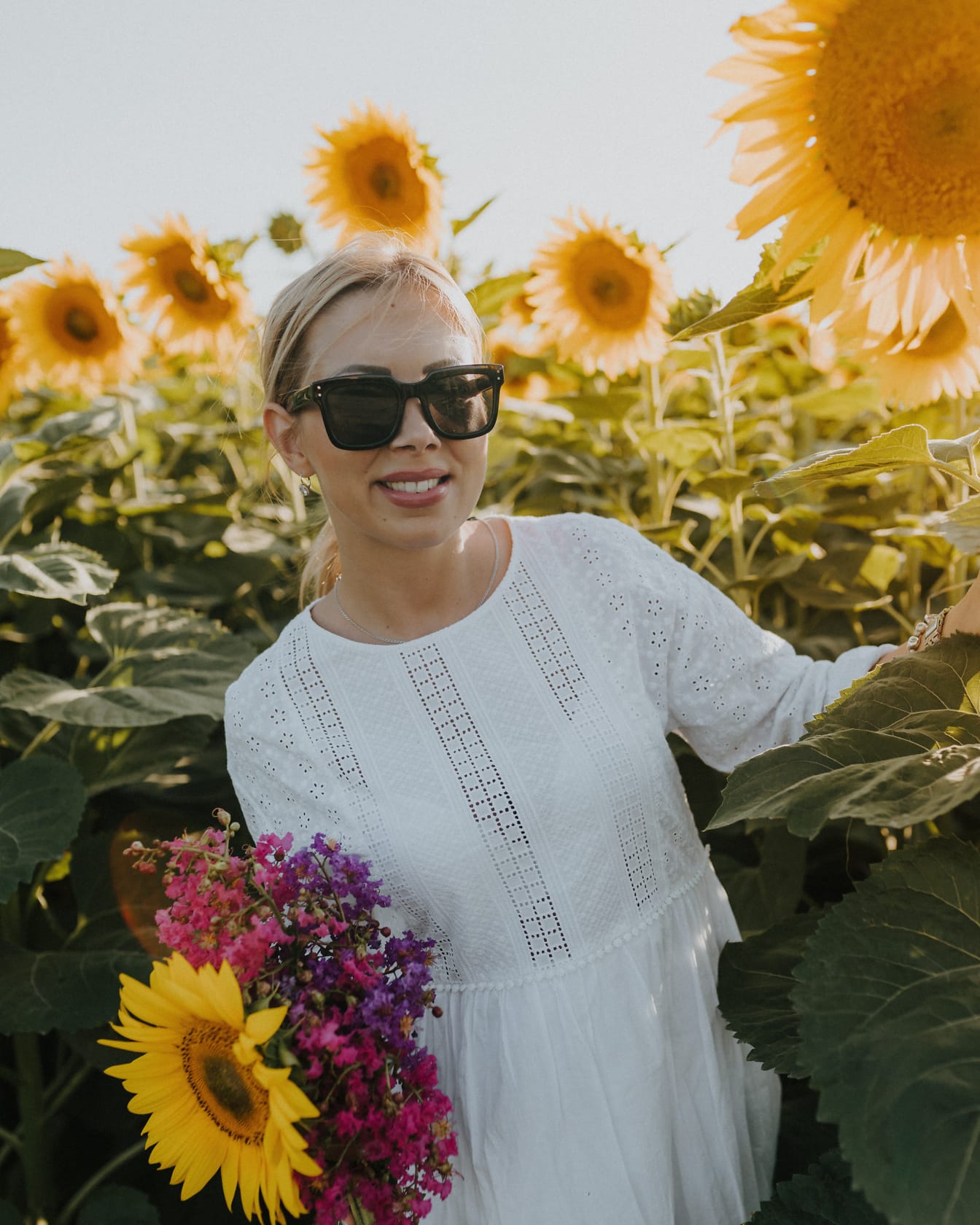 Leende blond kvinnafotomodell som bär solglasögon medan du poserar i ett fält av solrosor