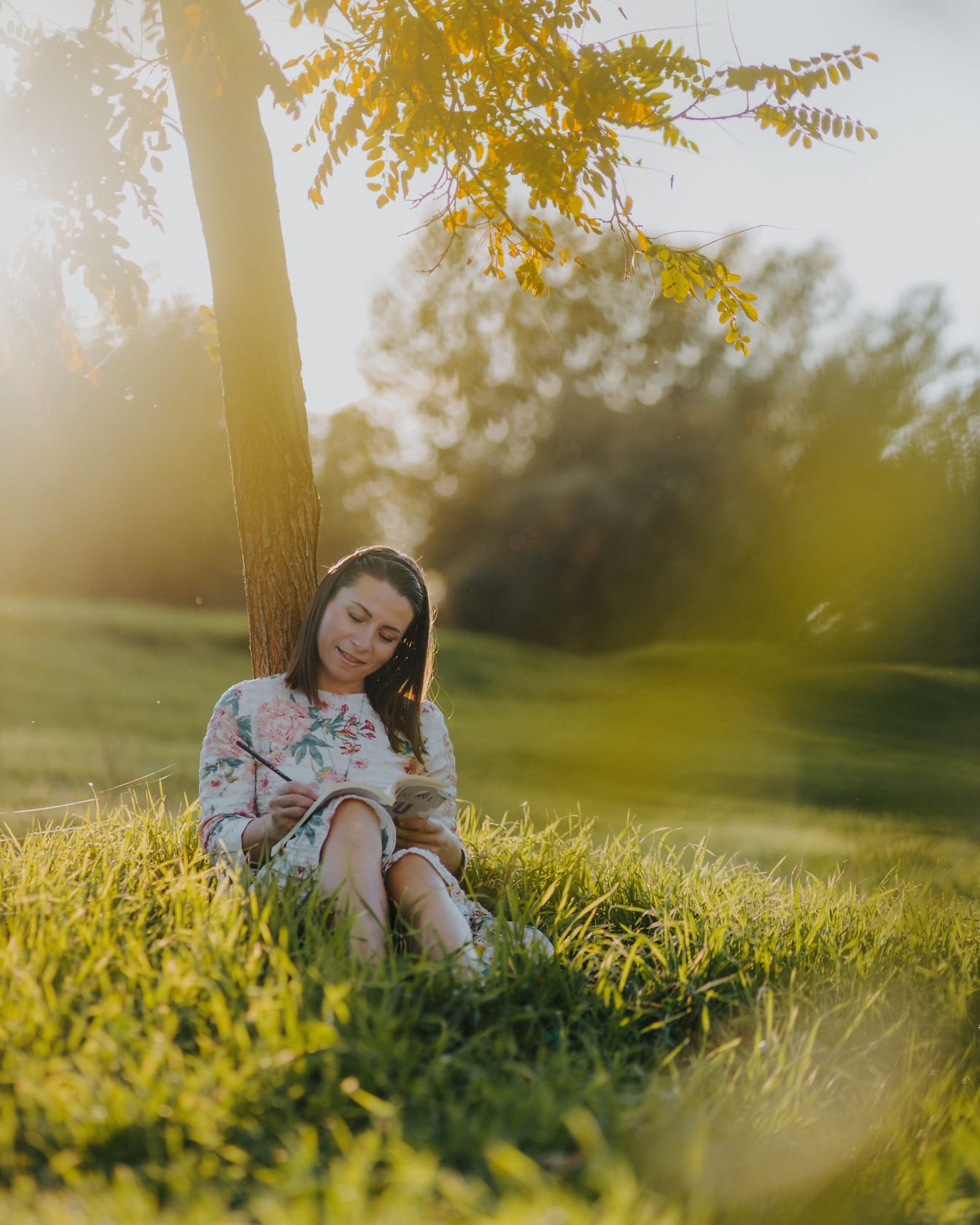 Seorang wanita tersenyum duduk di bawah pohon dan membaca buku pada hari musim panas yang cerah