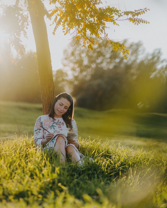 Une femme souriante est assise sous un arbre et lit un livre par une journée d’été ensoleillée
